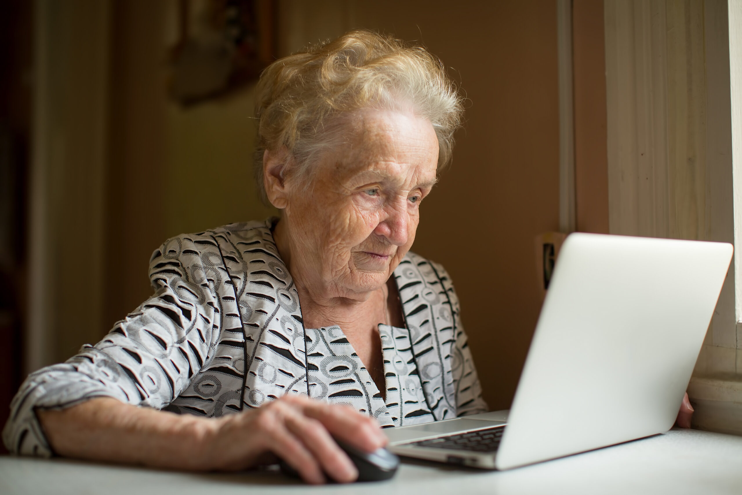 Eldre kvinne arbeider på bærbar PC. Sitter nær vinduet.